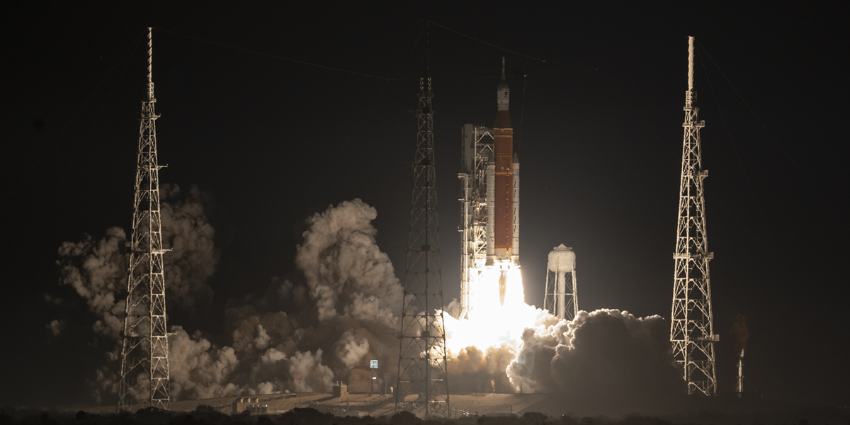 The Artemis I launch stage is illuminated against a black sky at Kennedy Space Center in Florida. NASA’s white Orion spacecraft is fixed to the top of the tall, cylindrical orange and white Space Launch System rocket. Bright yellow plumes are gushing from the bottom of the rocket as it begins its launch.