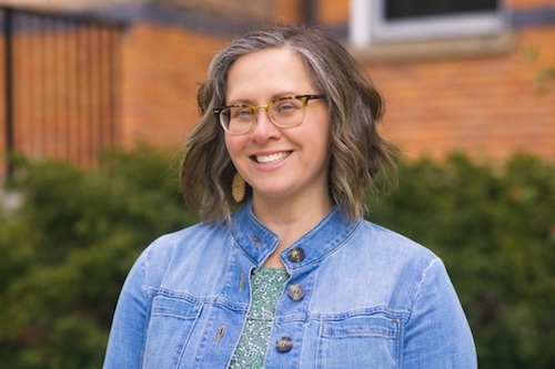 A headshot of Kristin Arola who is smiling and wearing a green shirt underneath a denim jacket.