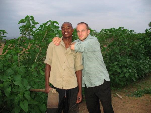 Two men posing for a picture in a crop field. One of them is holding a garden tool.