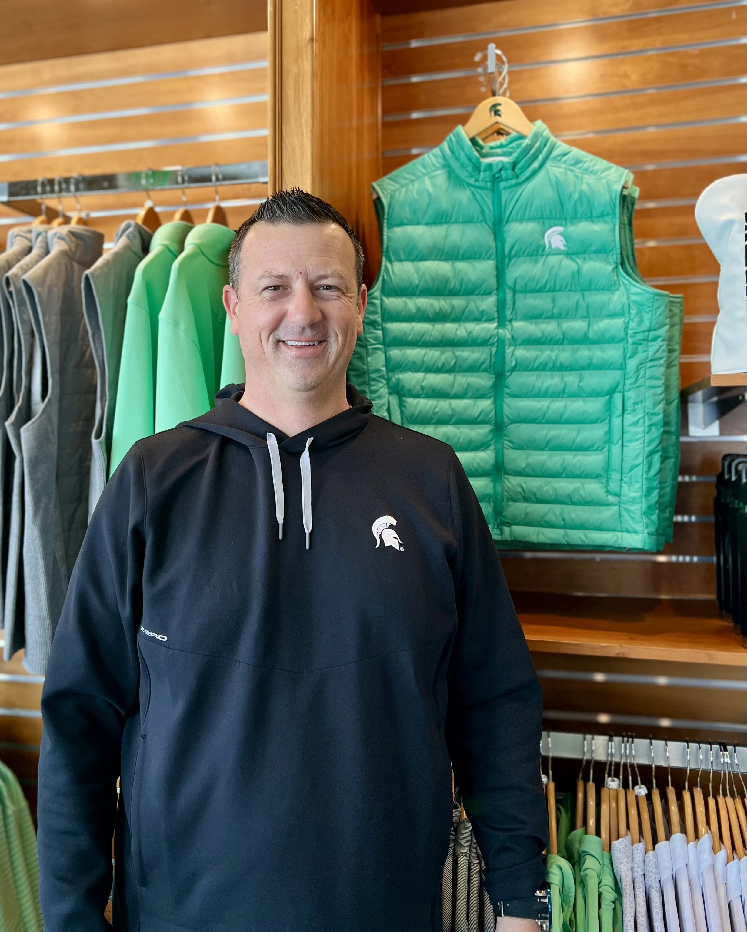 Bryan Harris wearing a black Michigan State University hoodie stands in front of green MSU apparel in a retail store.