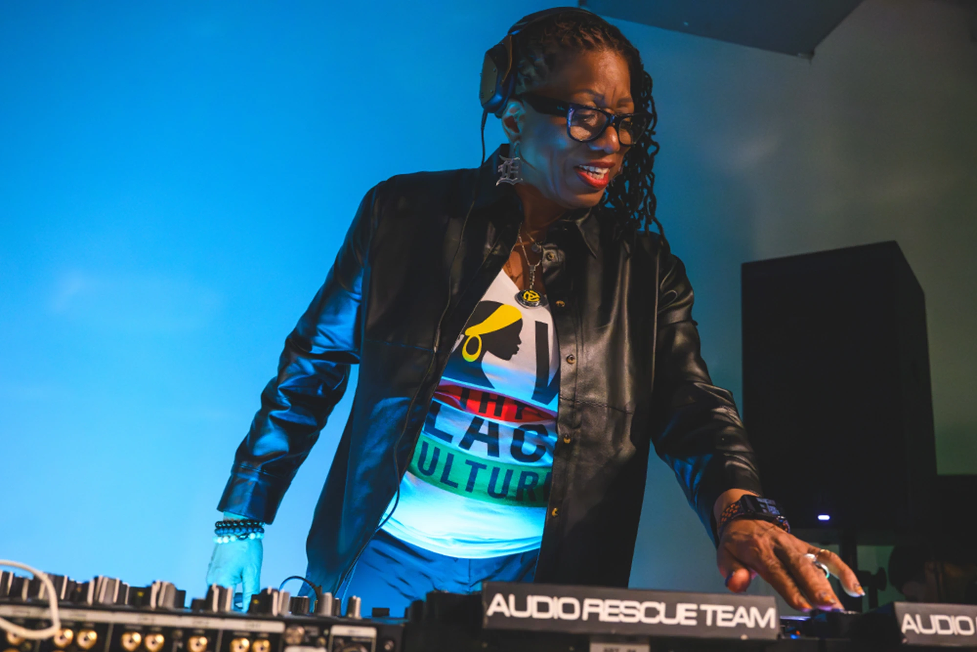 DJ Stacey “Hotwaxx” Hale wearing a black leather jacket and headphones mixes music behind a sound mixing board