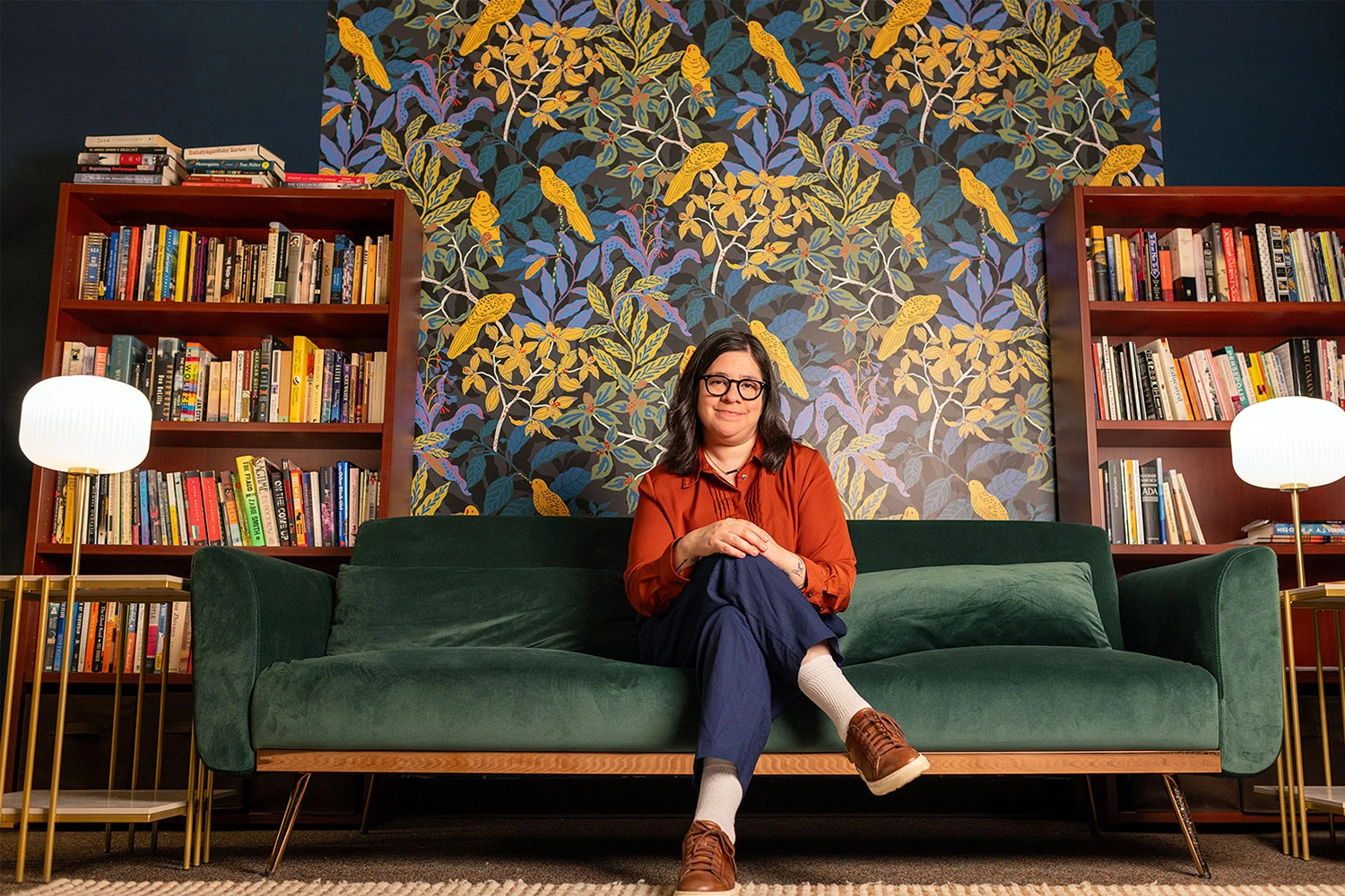 Alejandra Márquez, assistant professor of Spanish at Michigan State University, wears an orange shirt and glasses. She sits in a yellow chair and reads a book. A floor lamp is on the left side of the chair. A large indoor plant is on the right side of the chair.