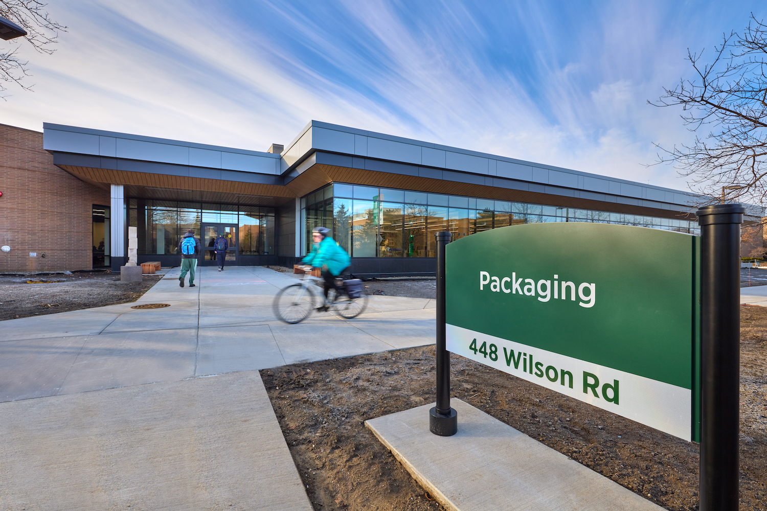 A biker rides in front of a modern looking building. A sign in front of the building reads: Packaging, 440 Wilson Rd
