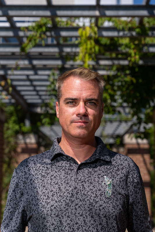 Rob Quinn in a gray Spartan golfing polo, standing beneath an arbor with greenery.