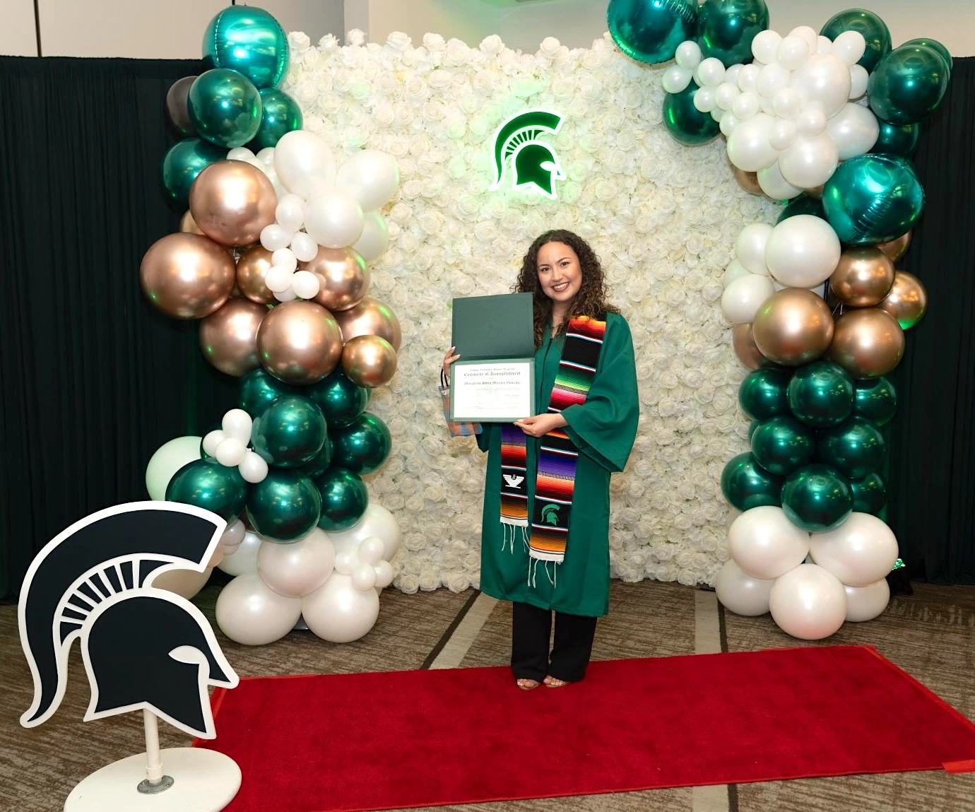 a student wearing a graduation gown, holding degree in front of a decorative green and white balloon backdrop